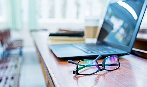 A pair of glasses on a table next to a laptop and coffee cup. 
